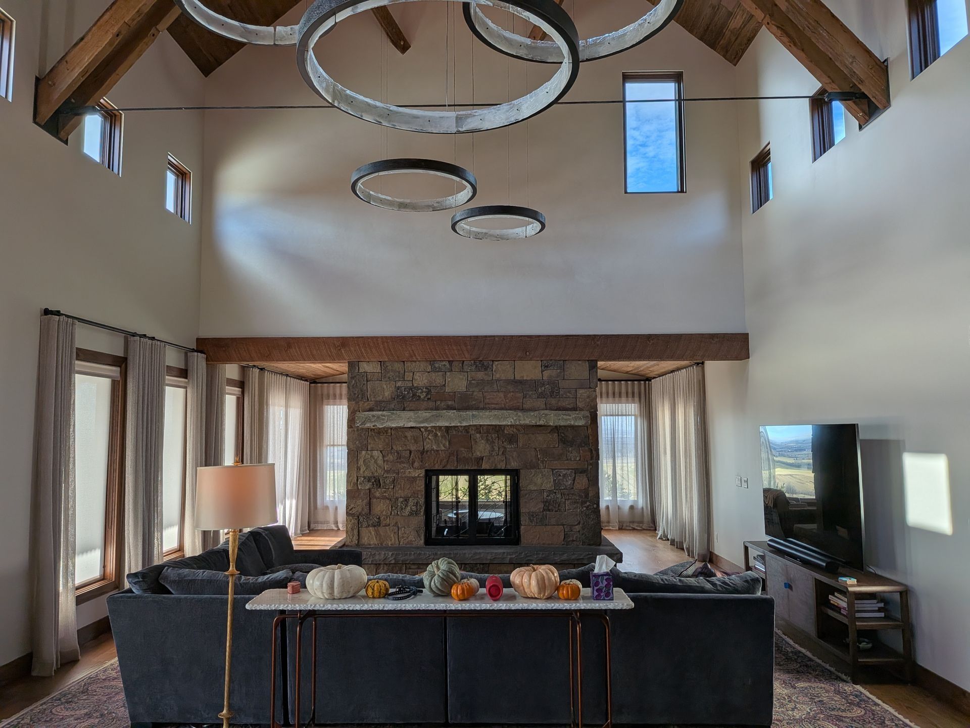 A spacious living room with high vaulted ceilings, a stone fireplace, ring chandeliers, and a sofa facing the hearth.