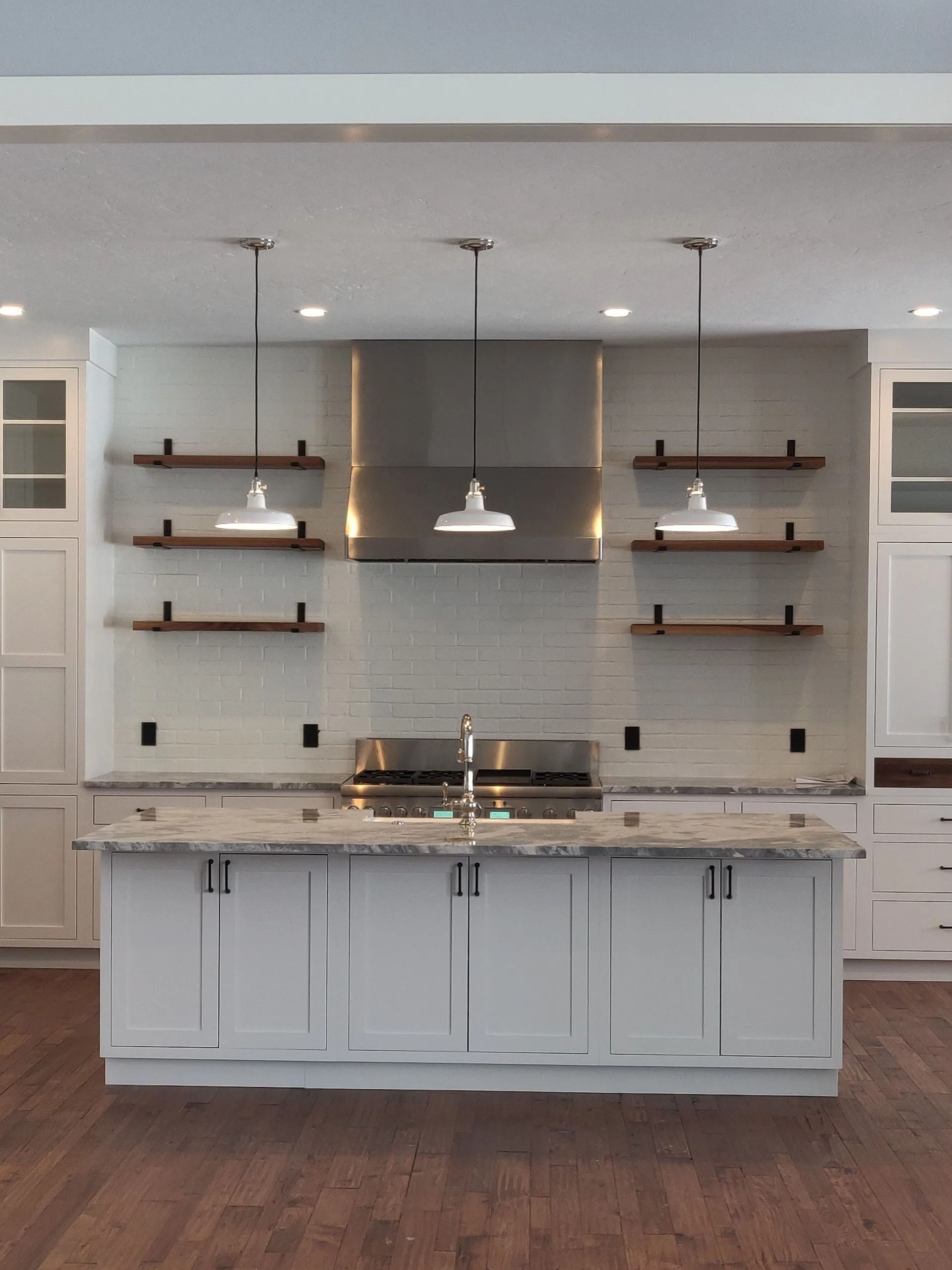 A kitchen island with a marble countertop sits in front of a white tiled backsplash, metal hood, and wood open shelving.