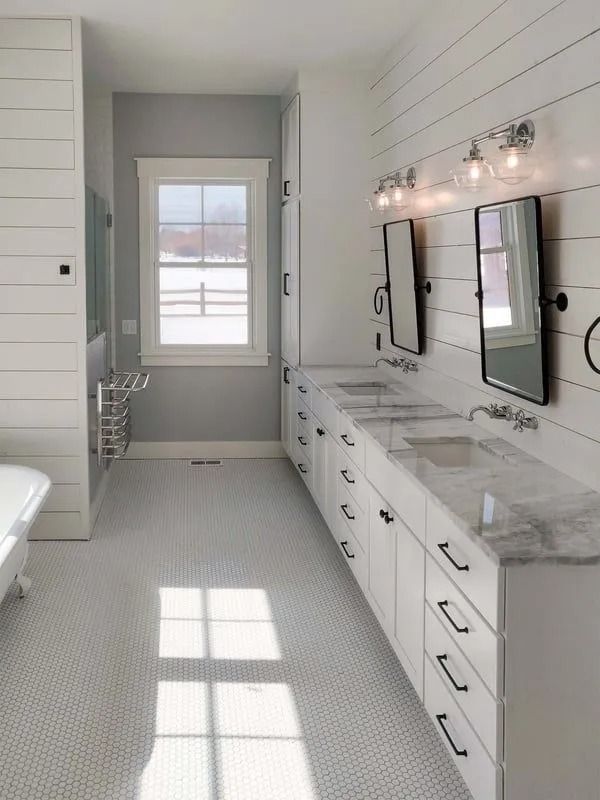 A bright bathroom with white shiplap walls, a double vanity with marble countertops, and a white tiled floor.