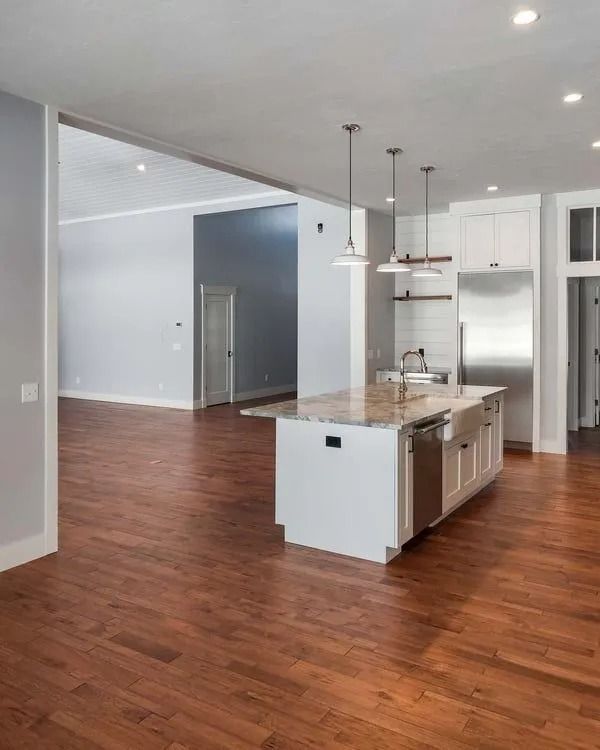 A modern kitchen with a white island, stainless appliances, and three pendant lights set against warm wood flooring.