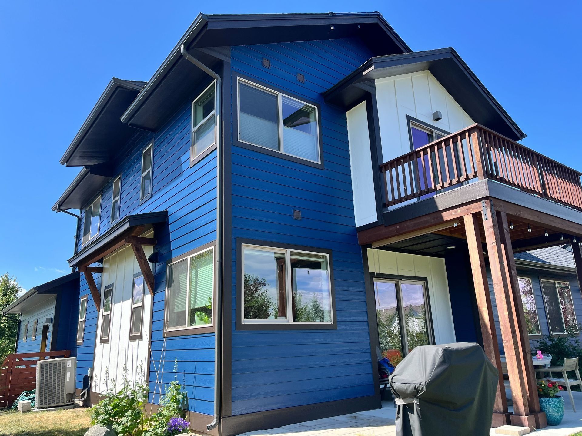 A two-story house with dark blue horizontal siding and white vertical accents, featuring a wooden balcony and patio.