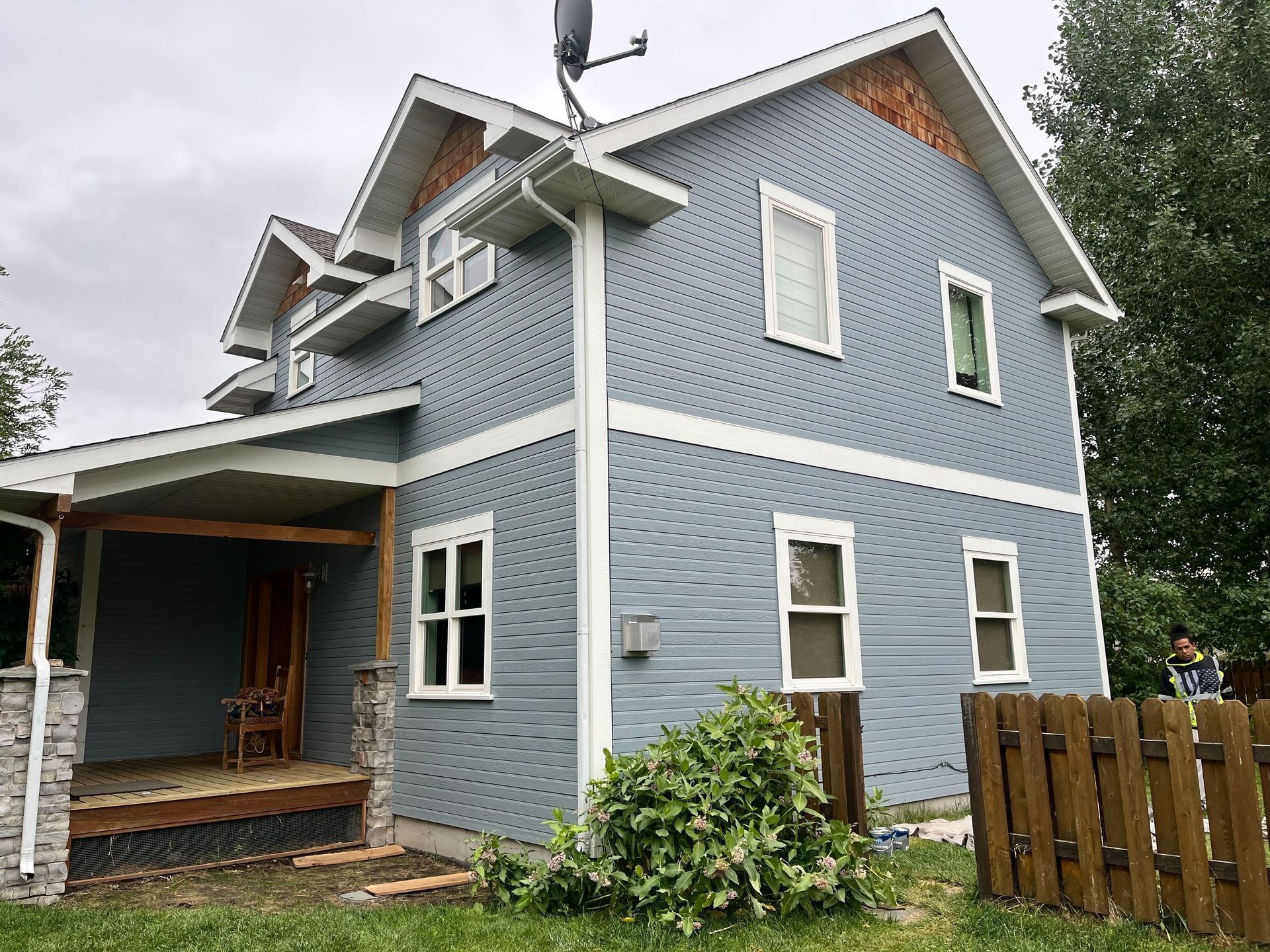 A two-story blue house with white trim and a covered front porch, seen from the yard next to a wooden fence.