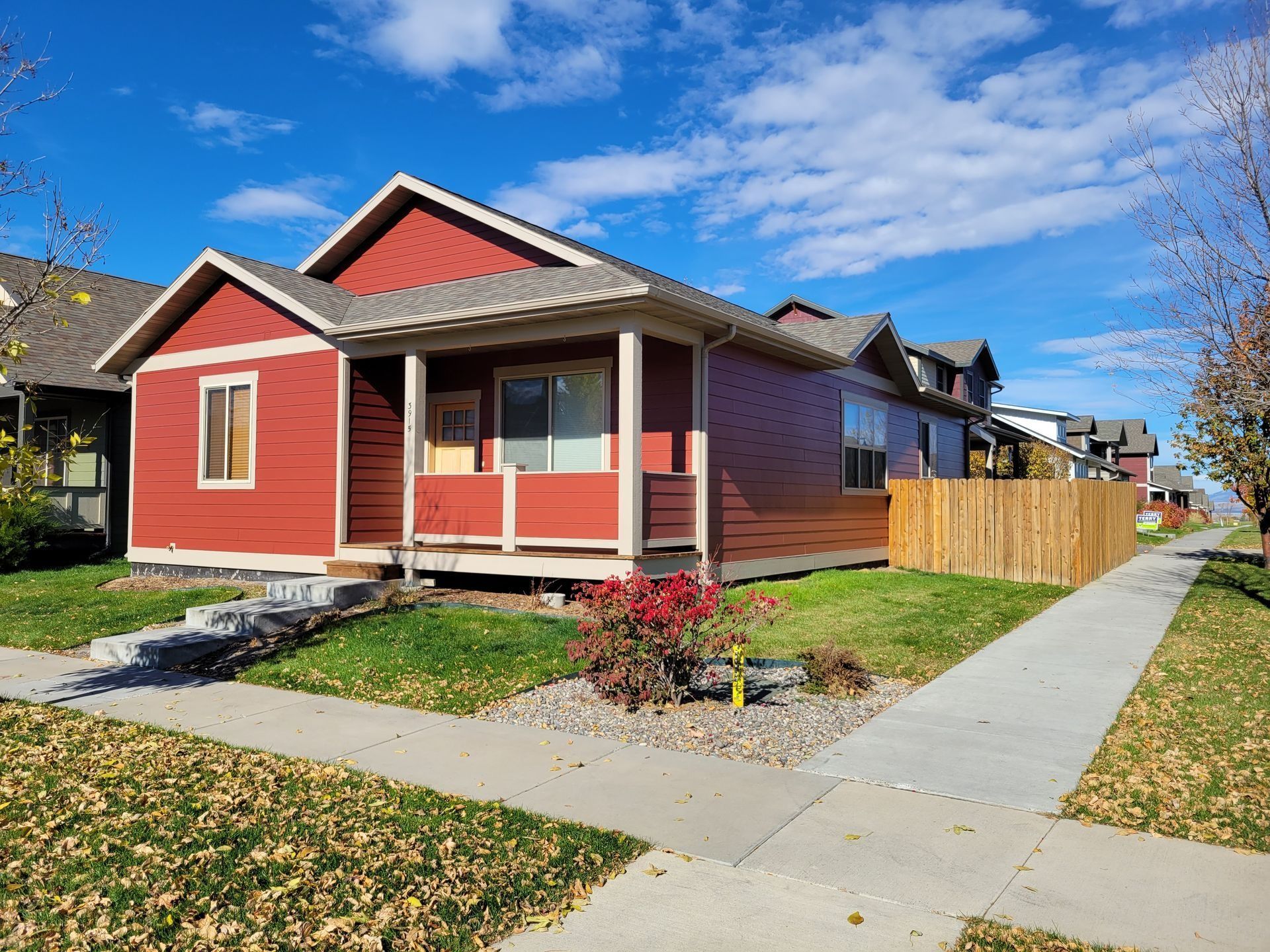 A red, single-story house with a front porch, tan trim, and a wooden fence, seen from a sidewalk on a sunny day.