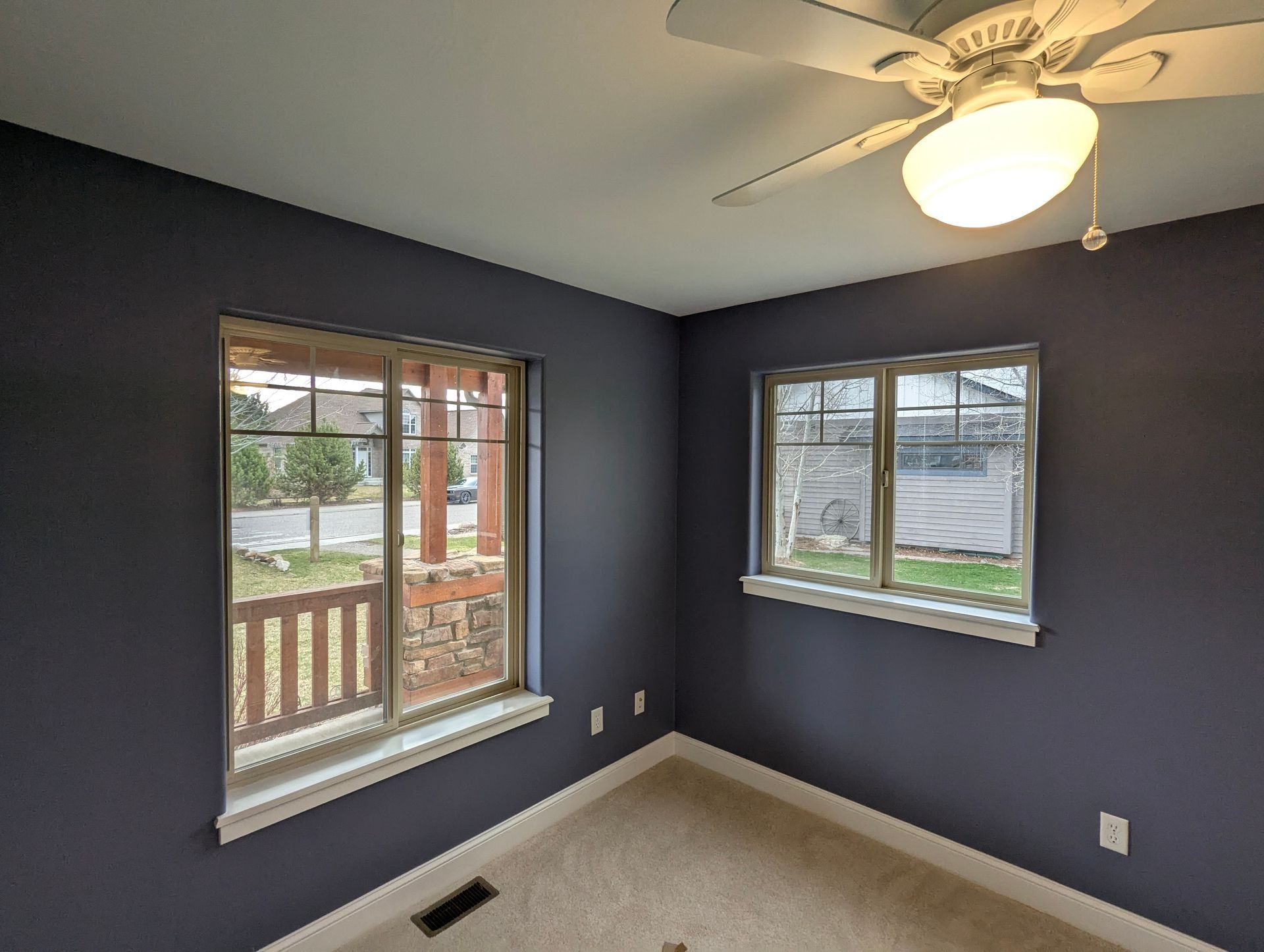 An empty room with dark blue walls, beige carpeting, two windows, and a ceiling fan.