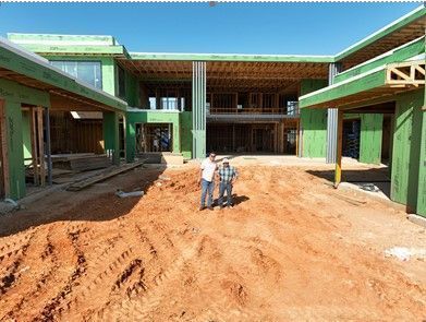 Two people stand on dirt ground in front of a large residential building under construction with green exterior panels.