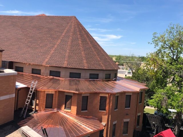 High-angle view of a building with a large brown shingled roof, a lower copper-trimmed roof, and a ladder against a wall.