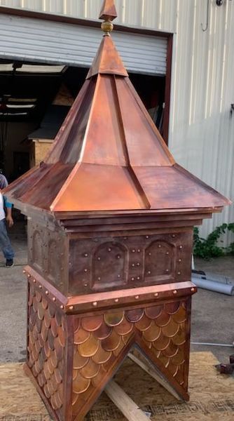 A copper cupola with a peaked roof, arched panels, and scalloped siding, sitting on a wooden platform in a workshop.