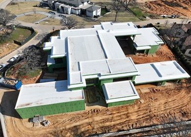 An aerial view of a construction site showing a modern house with a white flat roof and green plywood siding.