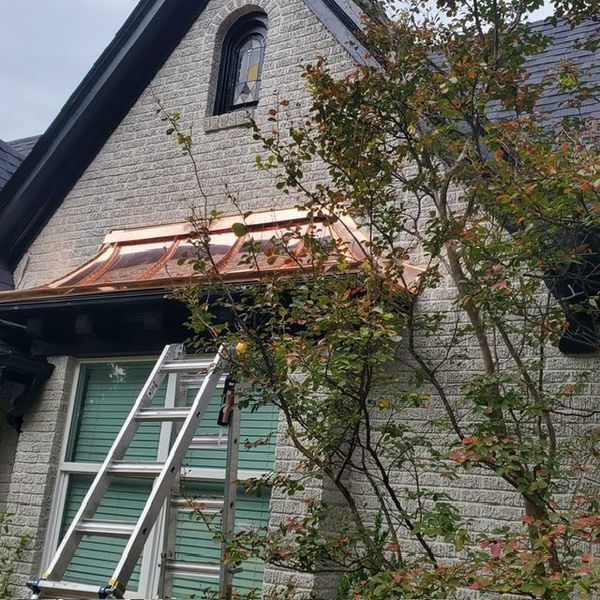 A metal ladder leans against a light-colored brick house wall with a new copper awning over a window.