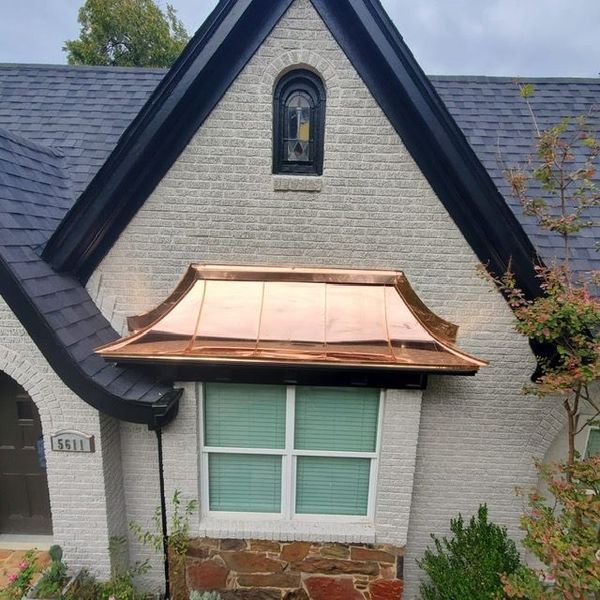 A stone house with a black shingled roof, a small arched window, and a new copper awning over a rectangular window.