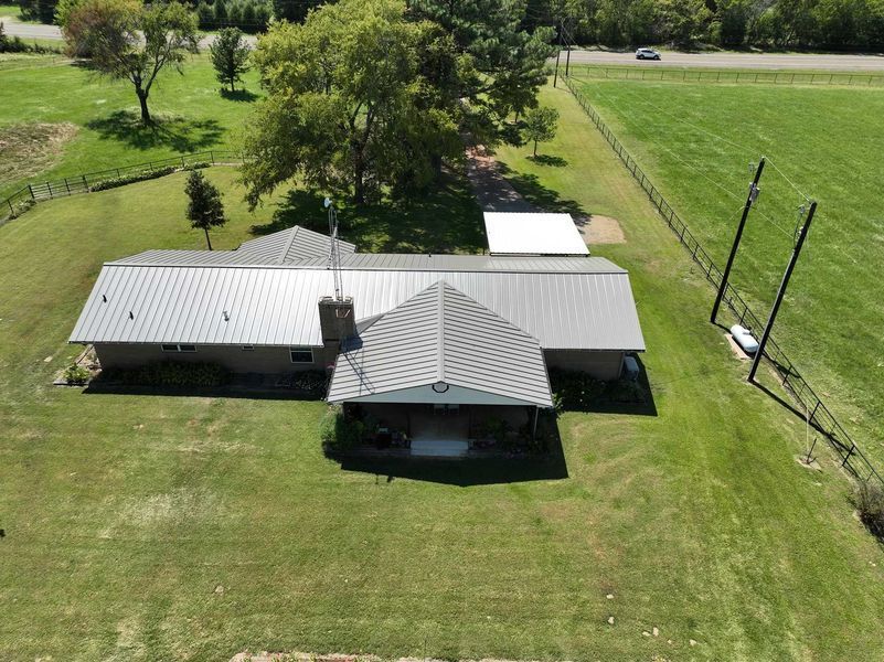 An aerial view of a single-story house with a metal roof and a prominent front porch, surrounded by a large green lawn.