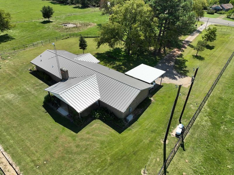 An aerial view of a brick house with a gray metal roof, a carport, and a surrounding grassy lawn with trees and a fence.