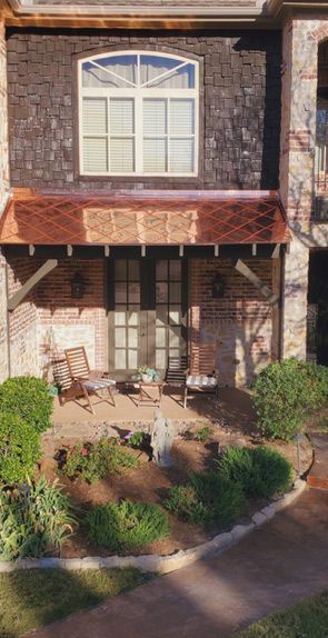A sunlit patio with two chairs and a statue, beneath a copper awning and a window on a stone-faced building.