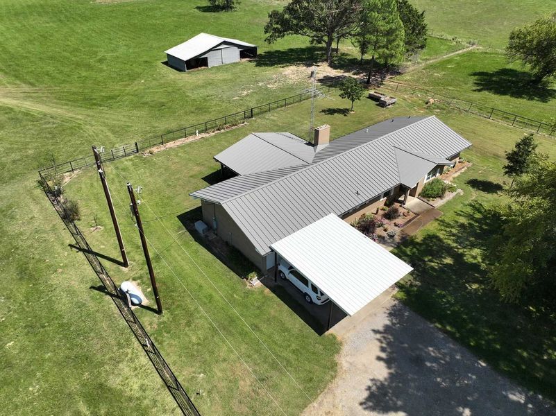 An aerial view of a ranch-style house with a metal roof, a carport, and a separate barn on a large, grassy lot.