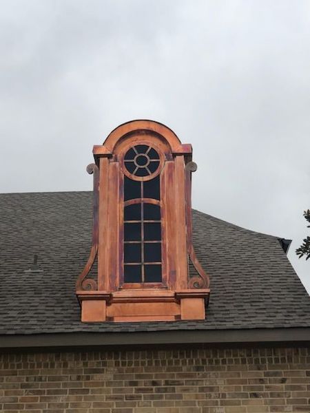 A decorative copper-clad arched dormer window set into a shingled residential roof above a brick wall.