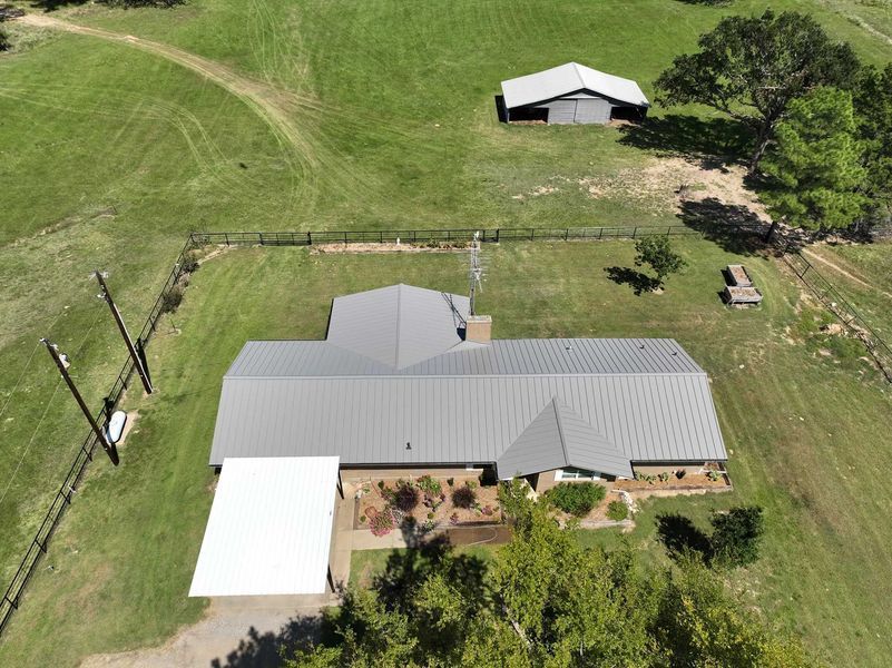 Aerial view of a single-story home with a grey metal roof, white carport, and a separate barn on a large grassy rural lot.