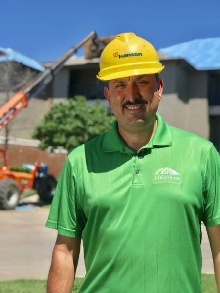A smiling person wearing a green polo shirt and a yellow construction hard hat stands in front of a building site.
