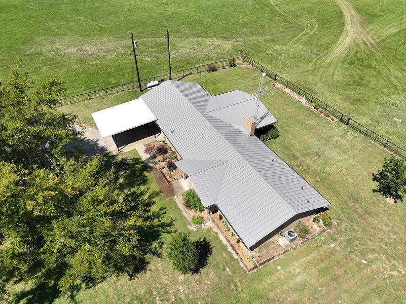 Aerial view of a rectangular house with a grey metal roof and white carport, situated on a grassy plot with a fence.