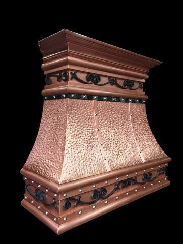 A hammered copper range hood with black iron scrolls and stud detailing, set against a black background.