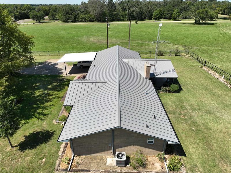 Aerial view of a house with a gray metal roof, a carport, and an antenna, surrounded by a large green field and trees.