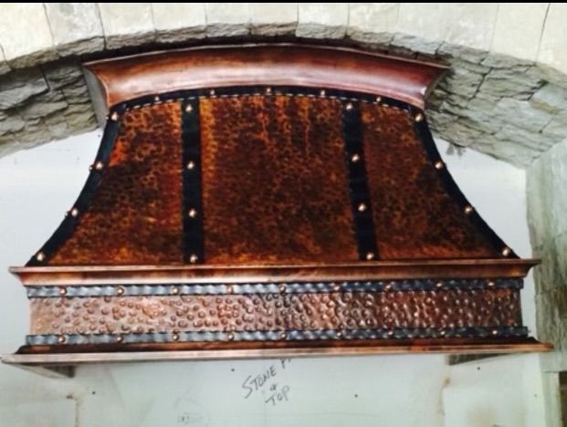 Ornate, hammered copper range hood with decorative dark metal straps and studs, mounted beneath a stone archway.