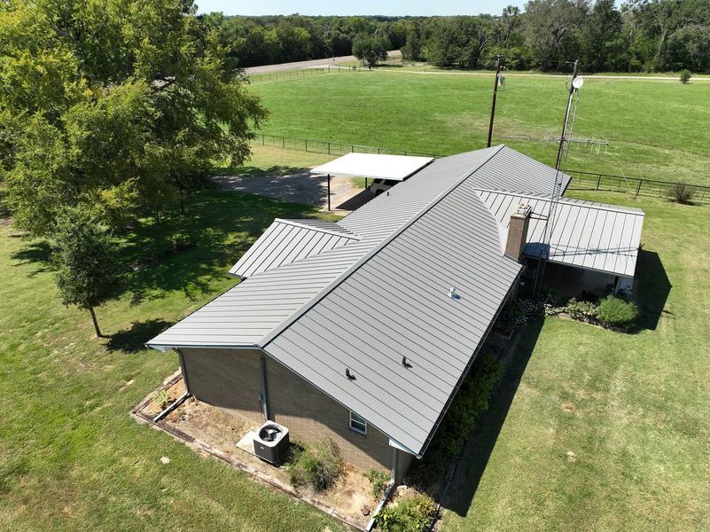 High-angle view of a brick house with a gray standing-seam metal roof, set in a rural landscape with trees and grass.