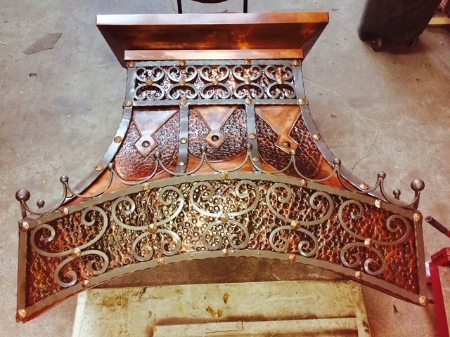 A custom hammered copper range hood with intricate wrought iron scrollwork overlays and decorative metal accents.