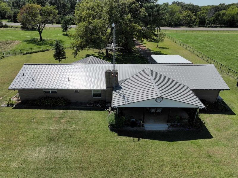 An aerial view of a single-story house with a light gray metal roof, a brick chimney, and a large covered patio area.