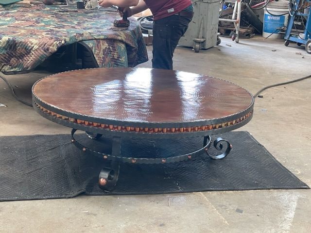 A round, hammered copper coffee table with a decorative metal trim and scrolled iron legs sits in a workshop.
