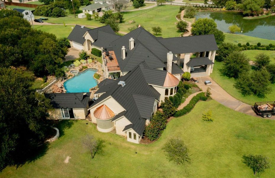 Aerial view of a large house with a dark roof and pool, situated on a lush green property with a pond.