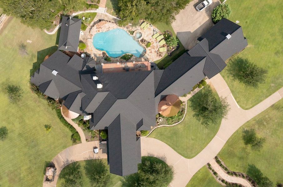 An aerial view of a large residential property featuring a complex dark roof, a swimming pool, and curved paved driveways.