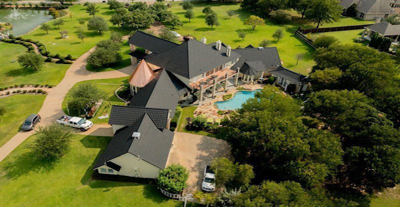 Aerial view of a sprawling luxury home with a dark roof, swimming pool, and fountain, surrounded by large, leafy trees.