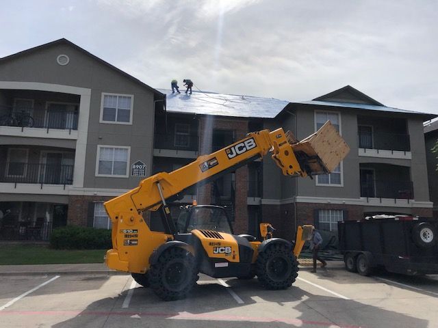 A yellow JCB telehandler lifts construction materials to two workers on the roof of a multi-story apartment building.