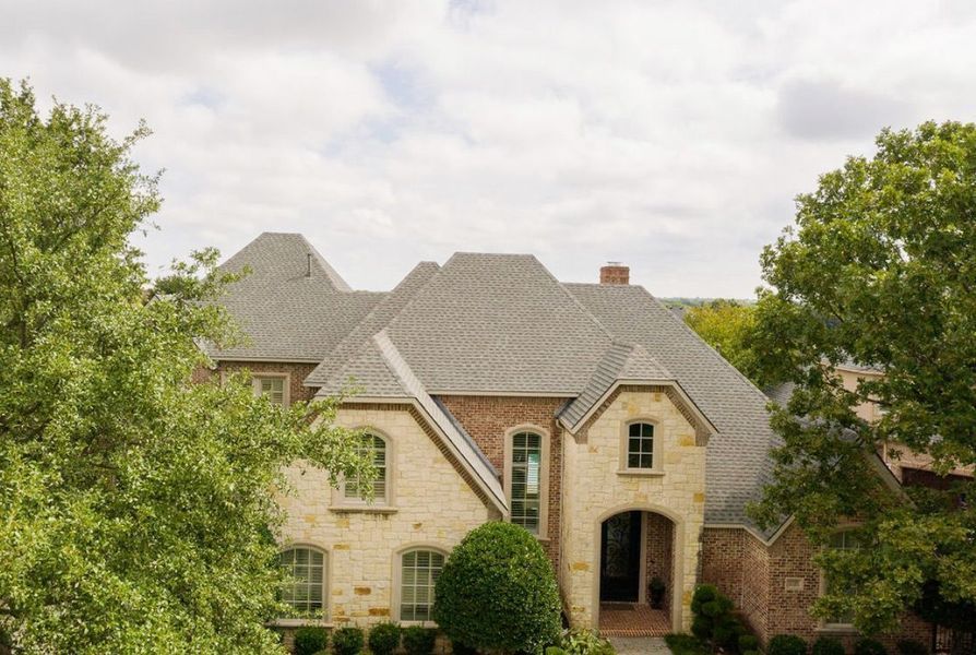 A two-story house with light-colored stone and red brick siding, a grey shingled roof, and green trees in front.