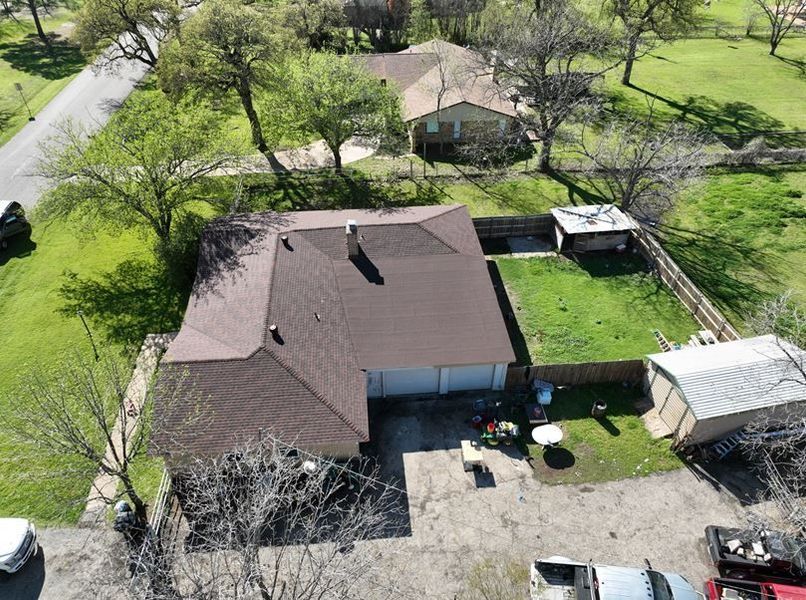 Aerial view of a residential house with a dark roof, a fenced backyard, and a gravel driveway with vehicles.