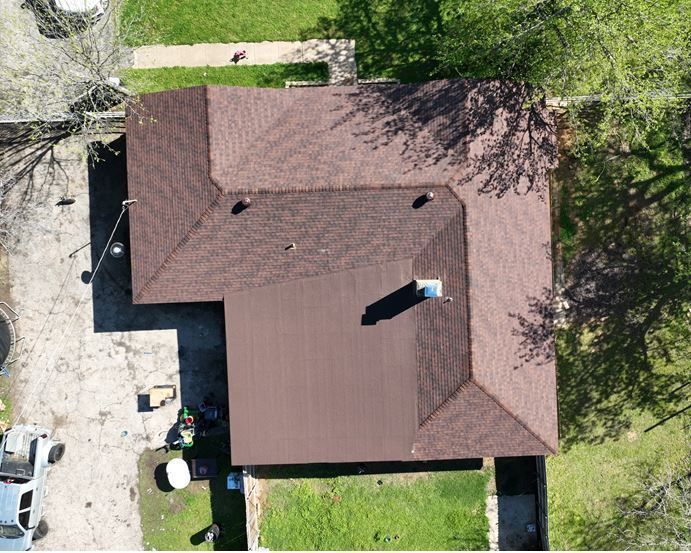 An aerial view of a brown shingled roof on an L-shaped house surrounded by trees and a driveway.