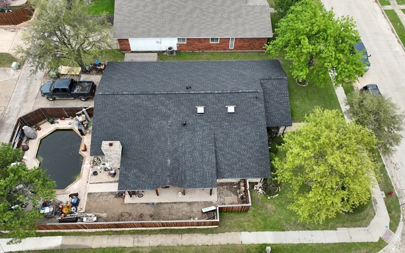 An aerial view of a dark-roofed suburban house with a backyard pool, stone chimney, and surrounding trees.