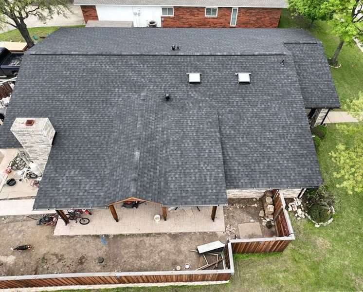 High-angle aerial view of a dark shingled roof with a stone chimney, covered patio, and a backyard area with a fence.