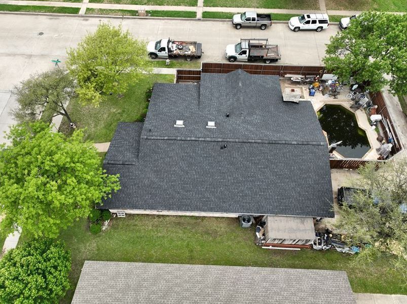 Aerial view of a dark shingled residential roof with a swimming pool, surrounding trees, and nearby street traffic.