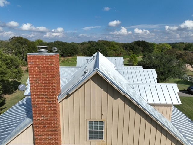 An aerial view of a tan house with a brick chimney and a silver metal roof, set against a green, wooded landscape.