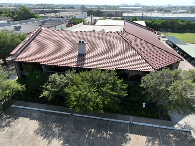 An aerial view of a building with a reddish-brown tile roof, a central chimney, and surrounding trees and parking lot.