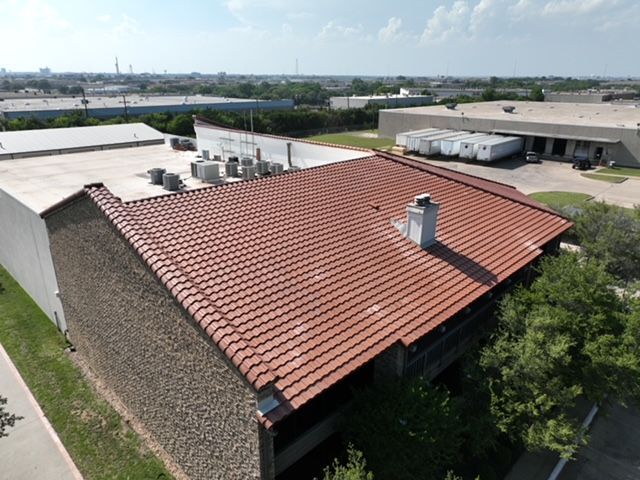 An aerial view of a building with a red tile roof and a chimney, situated next to a flat-roofed industrial warehouse.
