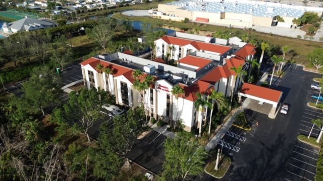Aerial view of a multi-story hotel with a U-shaped design, white walls, and a red tiled roof, surrounded by trees.