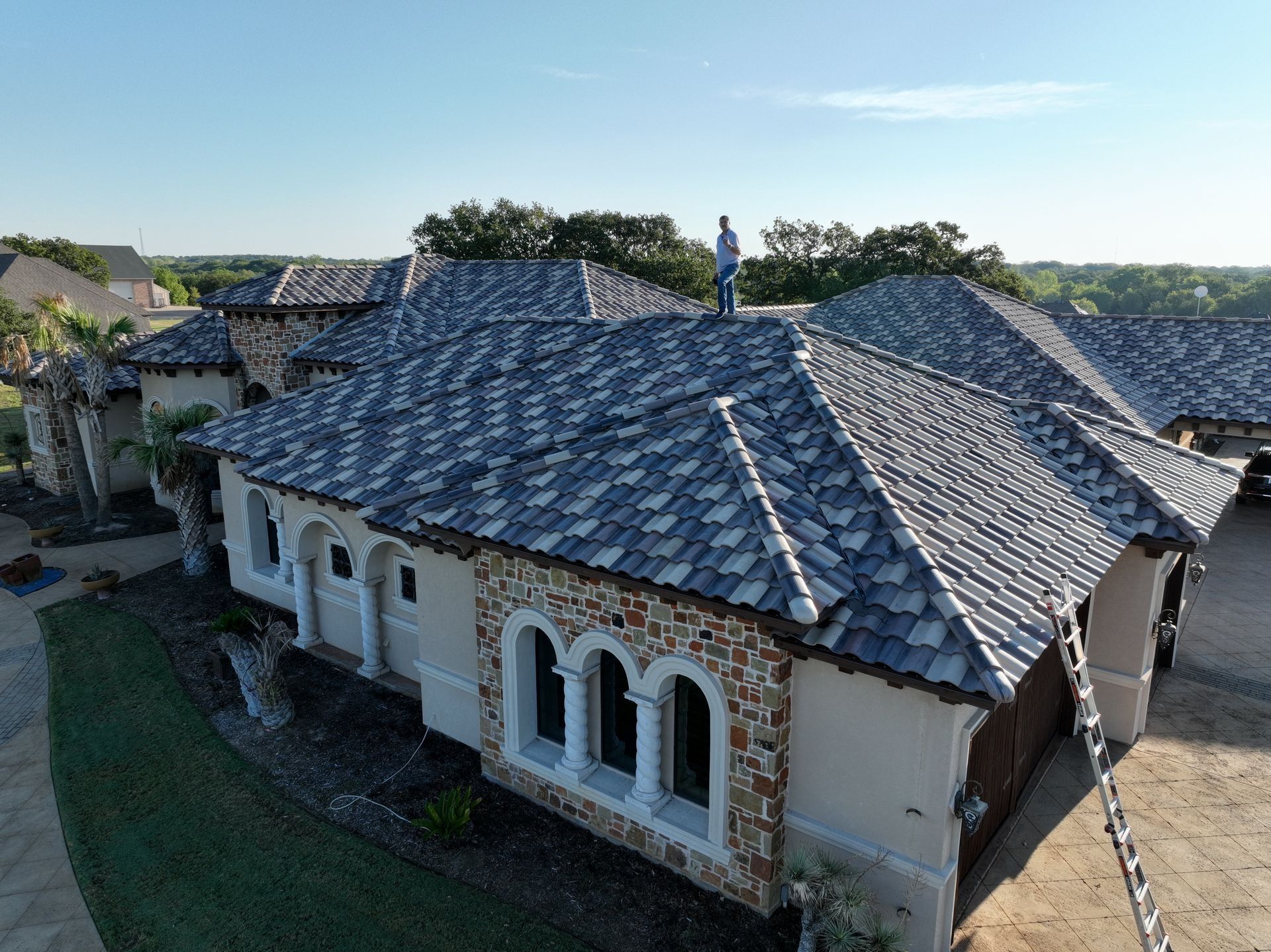 An aerial view of a person standing on the roof of a stone and stucco house with a dark, tiled roof and a ladder.