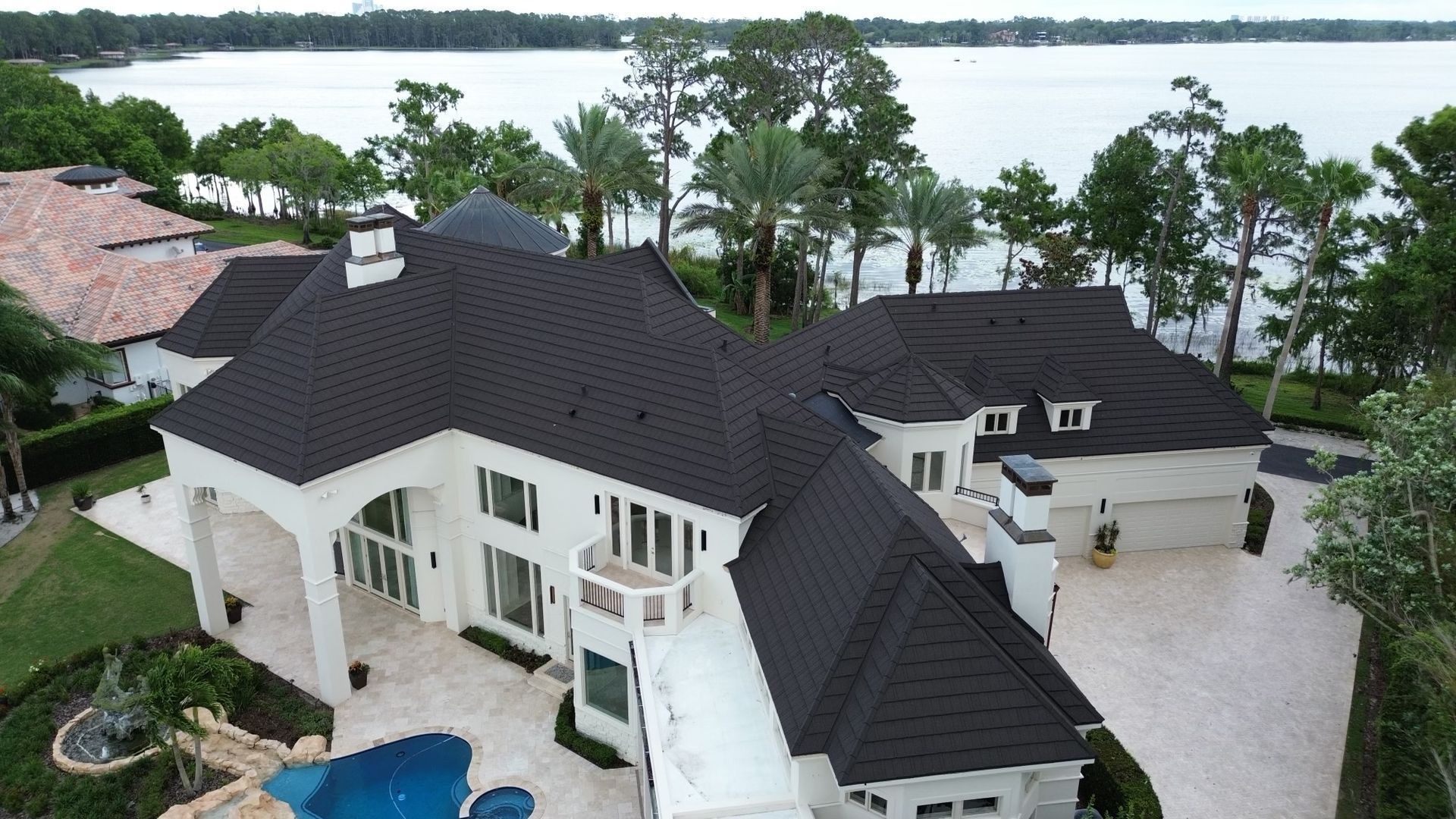 An aerial view of a white, multi-story luxury home with a dark roof, featuring a pool and lakefront backyard.