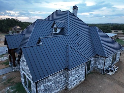 Aerial view of a house with a dark metal roof and stone exterior walls, surrounded by a rural landscape at dusk.