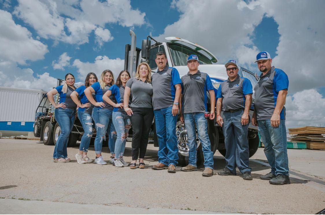 A group of people in company uniforms stand in a line in front of a semi-truck at an outdoor job site under a cloudy sky.