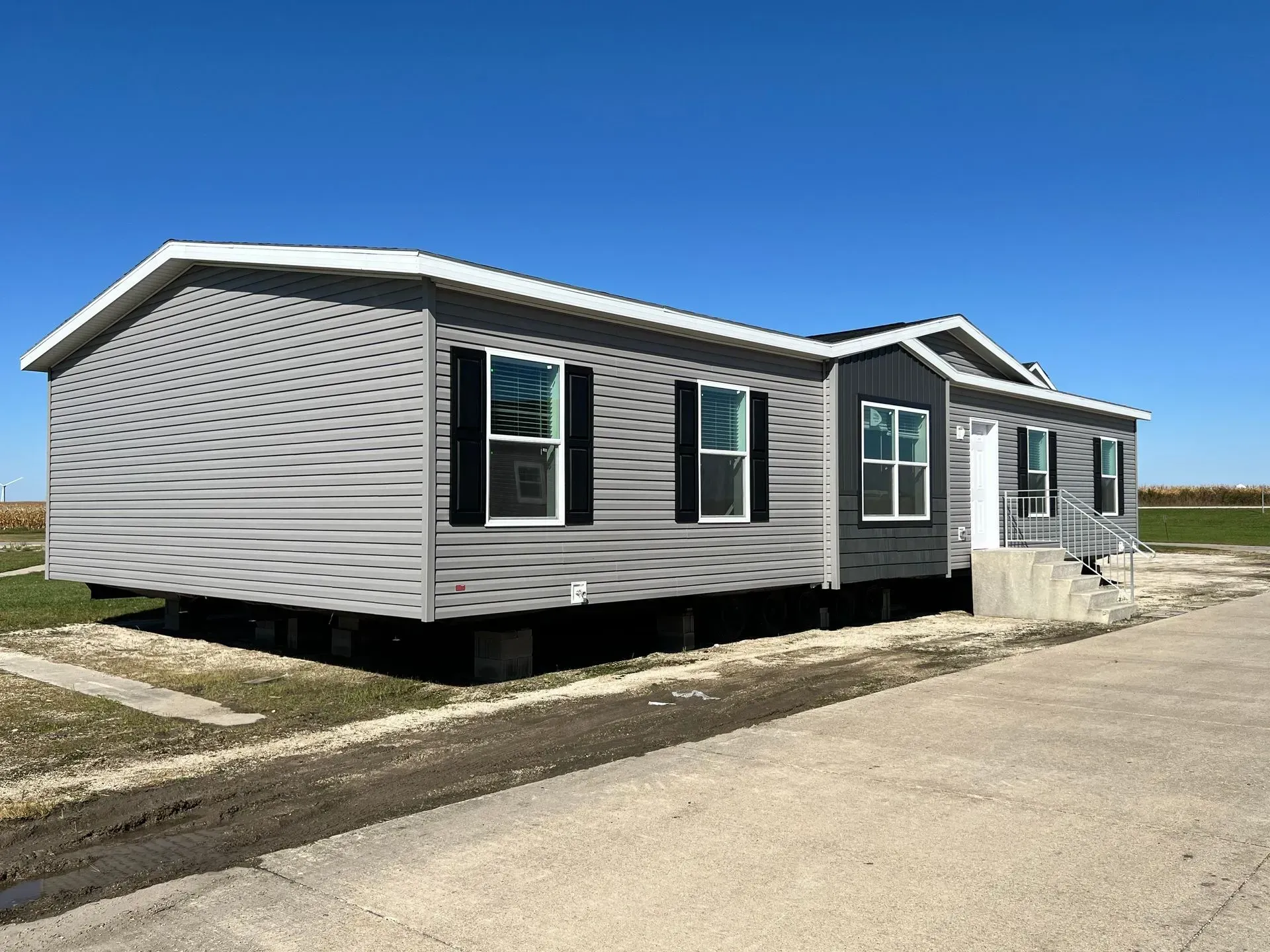 Gray single-story manufactured home with black shutters, set on a foundation, blue sky background.