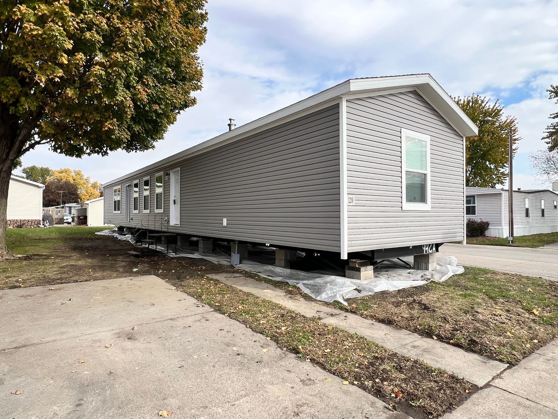 Gray mobile home on concrete blocks in a lot with nearby trees, overcast sky.
