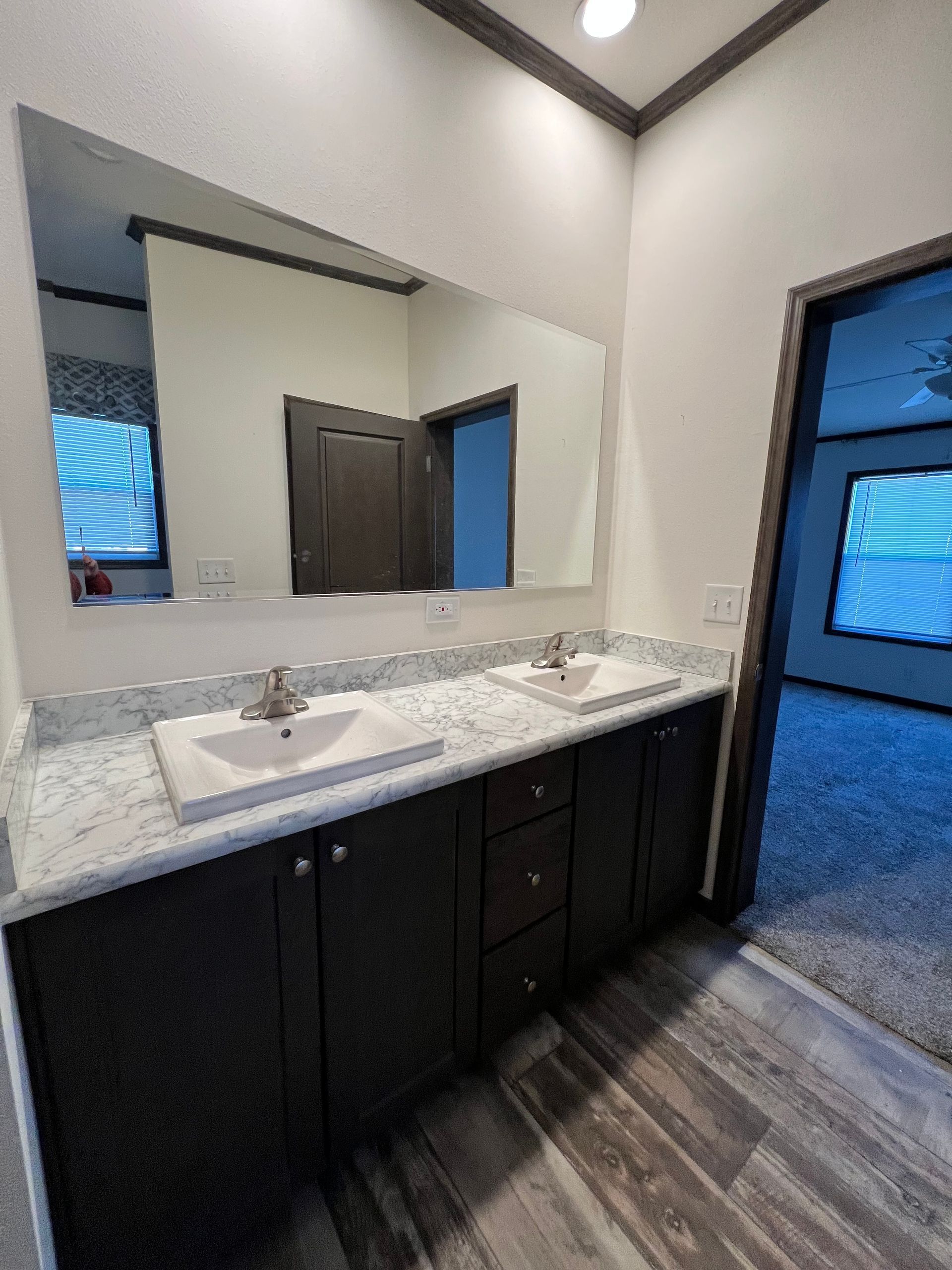 Bathroom with double sinks, dark cabinets, granite countertop, large mirror, and open doorway to bedroom.
